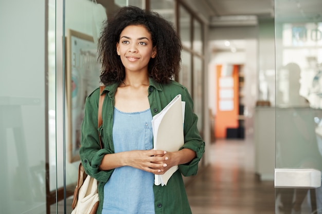 Ritratto di bella donna africana dello studente che sta nei libri sorridenti della tenuta del corridoio dell'istituto universitario che guardano nel lato. Concetto di educazione e apprendimento.