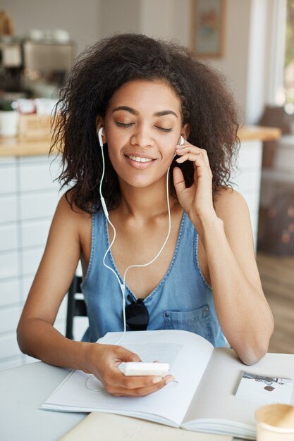 Ritratto di bella donna africana felice che ascolta la musica in seduta sorridente delle cuffie in caffè. Occhi chiusi.