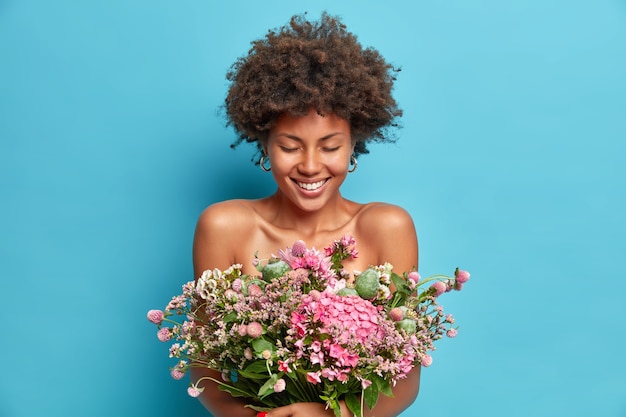 Ritratto di bella donna afroamericana dai capelli ricci pose con spalle nude ottiene presente dalla persona amata gode di primavera ha umore felice isolato sopra la parete blu dello studio