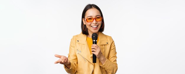 Ritratto di bella donna asiatica in occhiali da sole ragazza alla moda che canta dando discorso con microfono tenendo il microfono e sorridente in piedi su sfondo bianco Copia spazio