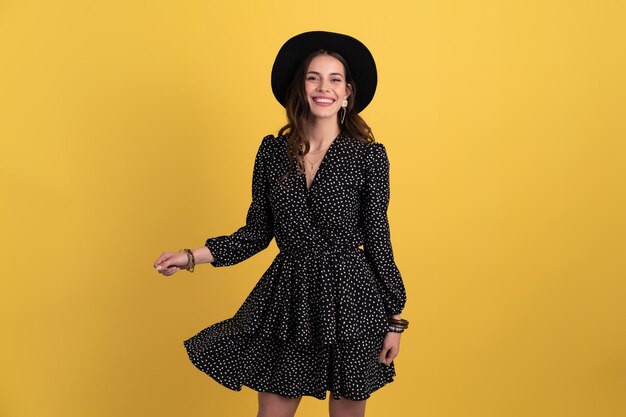 Ritratto di bella donna attraente in posa isolato su sfondo giallo che indossa abito nero punteggiato e cappello nero alla moda primavera estate accessori stile moda sorridente umore felice divertendosi