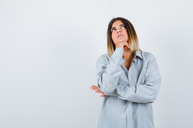 Ritratto di bella donna che sostiene il mento a portata di mano, alzando lo sguardo in camicia e guardando la vista frontale pensierosa