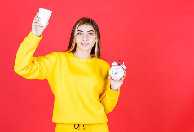 Ritratto di bella donna che tiene tazza e orologio sulla parete rossa