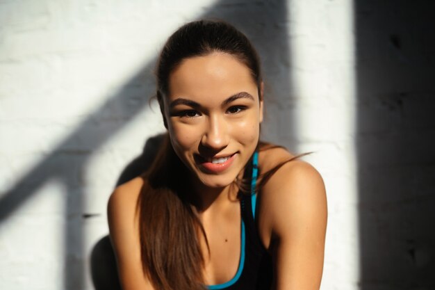 Ritratto di bella donna. Chiuda sul fronte della donna sportiva che esamina macchina fotografica sulla parete grigia con lo spazio della copia. Felice ragazza in forma in abbigliamento sportivo che ride