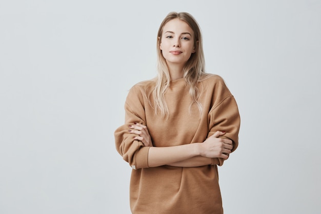 Ritratto di bella donna con i capelli lisci biondi che sorride allegramente mentre ascolto complimenti, indossa maglione a maniche lunghe sciolto, tenendo le braccia conserte.