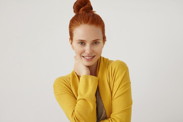 Ritratto di bella donna con i capelli rossi panino, pelle lentigginosa, tenendo la mano sul collo, indossa un maglione giallo, guardando con fiducia e felicemente