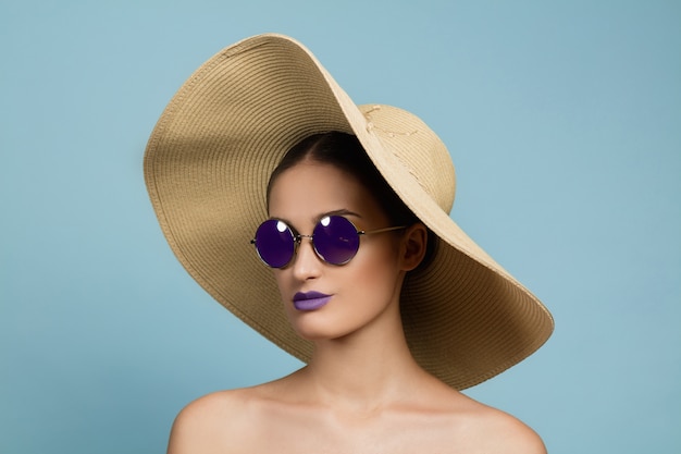 Ritratto di bella donna con trucco luminoso, cappello e occhiali da sole in studio blu