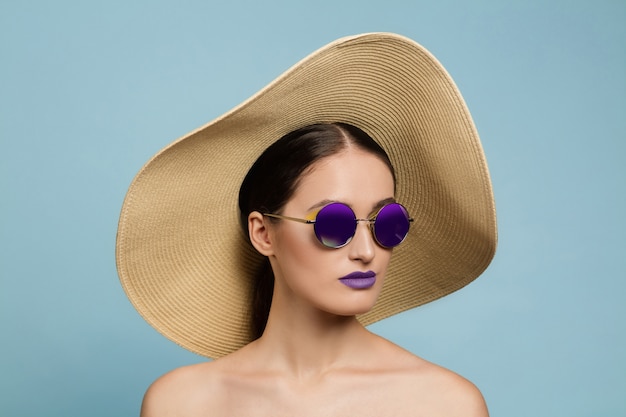 Ritratto di bella donna con trucco luminoso, cappello e occhiali da sole su sfondo blu studio. Trucco e acconciatura alla moda e alla moda. Colori dell'estate. Concetto di bellezza, moda e annuncio. Grave.