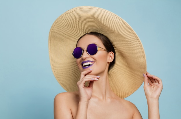 Ritratto di bella donna con trucco luminoso, cappello e occhiali da sole su sfondo blu studio. Trucco e acconciatura alla moda e alla moda. Colori dell'estate. Concetto di bellezza, moda e annuncio. Ridendo.