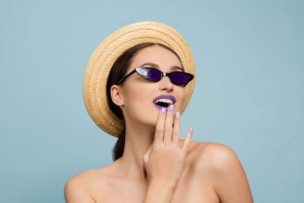 Ritratto di bella donna con trucco luminoso, cappello e occhiali da sole su sfondo blu studio. Trucco e acconciatura alla moda e alla moda. Colori dell'estate. Concetto di bellezza, moda e annuncio. Stupito.