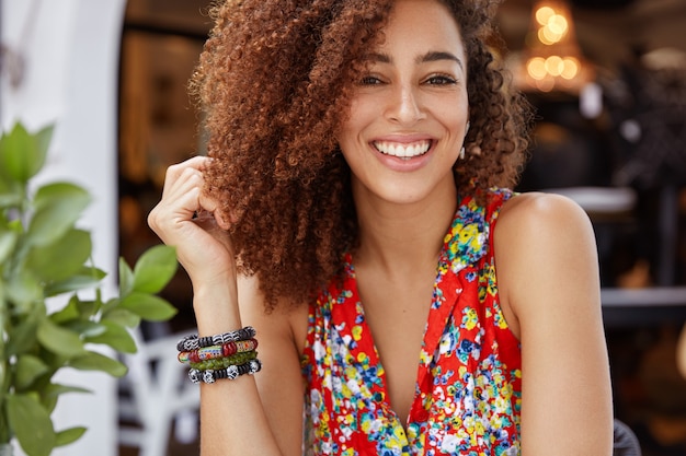 Ritratto di bella donna dalla pelle scura felice con i capelli ricci e un ampio sorriso brillante, dimostra emozioni positive, indossa elegante camicetta luminosa.