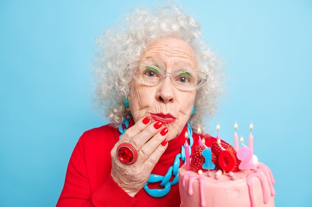 Ritratto di bella donna di bell'aspetto indossa gioielli e abiti rossi festivi tiene la torta con le candele festeggia il 102esimo compleanno essendo in festa per gli anziani