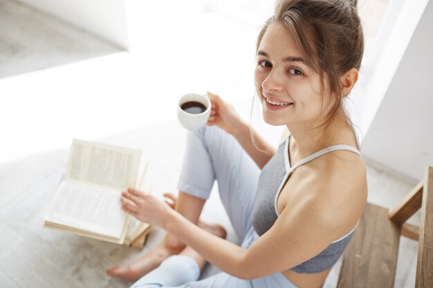 Ritratto di bella donna felice sorridente tenendo la tazza di caffè seduto sul pavimento con il libro.