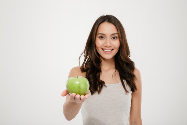 Ritratto di bella donna in buona salute che sorride e che mostra mela succosa verde sulla macchina fotografica, isolata sopra bianco