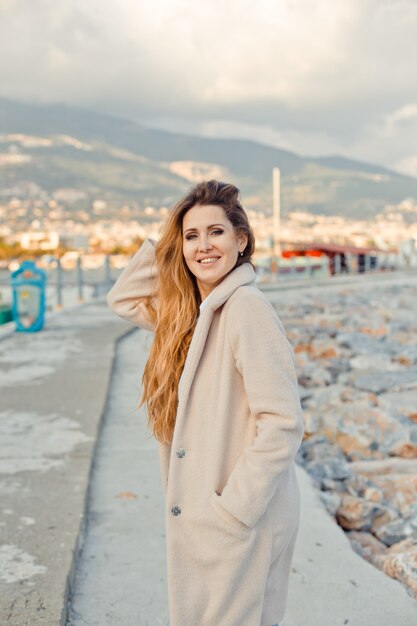 Ritratto di bella donna in piedi, tenendo i capelli e sorridendo in riva al mare durante il giorno.