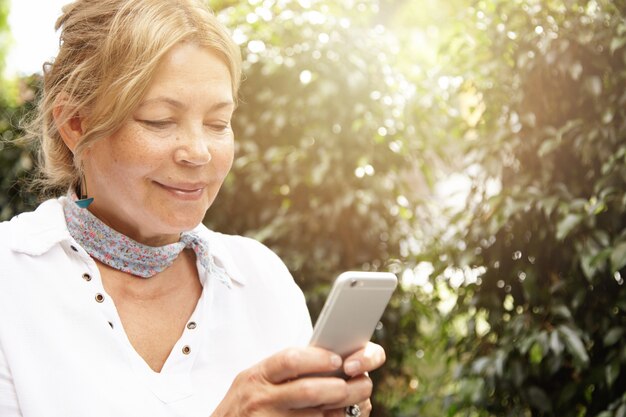 Ritratto di bella donna matura con capelli chiari utilizzando lo smartphone, digitando i messaggi tramite i social network mentre era seduto nel suo giardino nel giorno di sole, sorridendo mentre chattava con i suoi figli