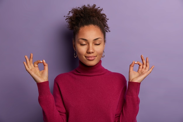 Ritratto di bella donna medita, tiene entrambe le mani nel gesto giusto, tiene gli occhi chiusi, pratica yoga per rilassarsi dopo il lavoro