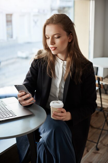 Ritratto di bella donna moderna nella caffetteria, seduto vicino alla finestra, bere caffè e navigazione in smartphone, avendo una pausa di lavoro con il computer portatile