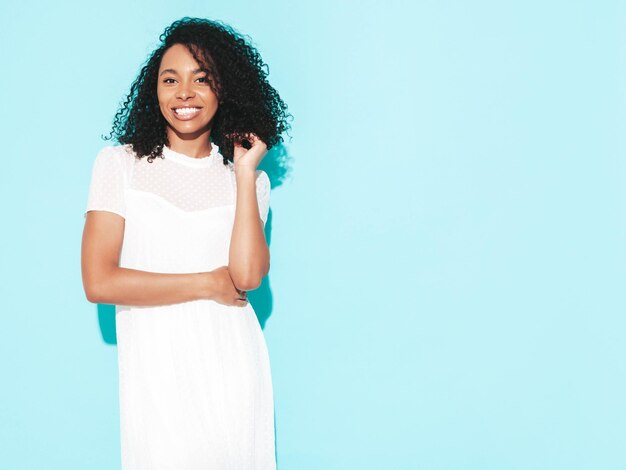 Ritratto di bella donna nera con l'acconciatura afro riccioli Modello sorridente vestito in abito estivo bianco Femmina spensierata sexy in posa vicino al muro blu in studio Abbronzato e allegro Isolato