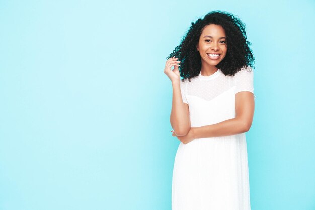 Ritratto di bella donna nera con l'acconciatura afro riccioli Modello sorridente vestito in abito estivo bianco Femmina spensierata sexy in posa vicino al muro blu in studio Abbronzato e allegro Isolato