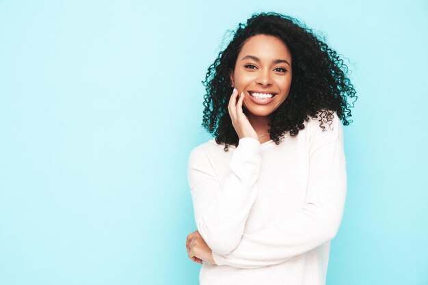 Ritratto di bella donna nera con l'acconciatura afro riccioli Modello sorridente vestito in abito estivo bianco Femmina spensierata sexy in posa vicino al muro blu in studio Abbronzato e allegro Isolato