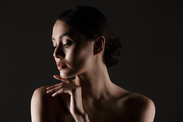 Ritratto di bella donna sensuale che osserva da parte mentre si tocca il mento in condizioni di scarsa luminosità, isolato sopra il nero