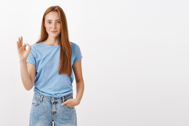 Ritratto di bella donna soddisfatta e felice della testarossa in maglietta blu che tiene la mano in tasca che mostra il gesto giusto o perfetto con le dita che sorridono gradimento o che accettano il piano