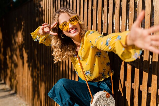 Ritratto di bella donna sorridente alla moda che ride emotiva bionda in camicetta gialla che indossa occhiali da sole, borsa di paglia stile bali