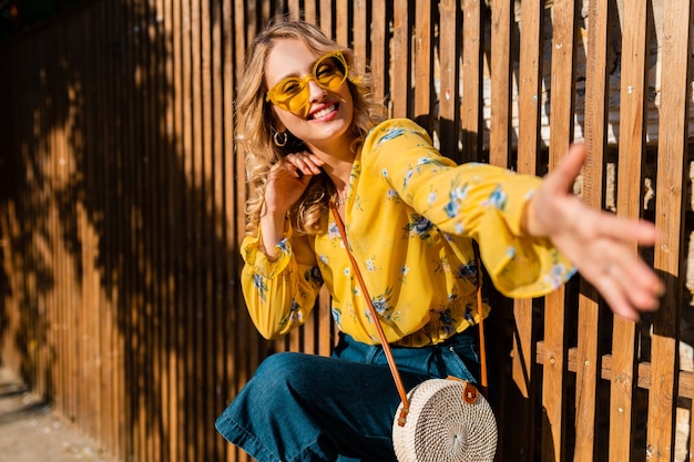 Ritratto di bella donna sorridente alla moda che ride emotiva bionda in camicetta gialla che indossa occhiali da sole, borsa di paglia stile bali