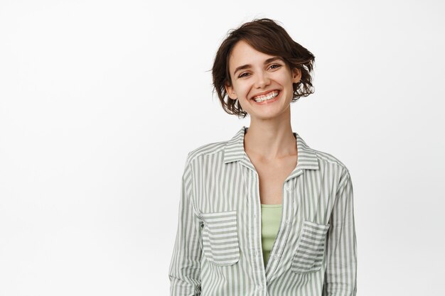 Ritratto di bella donna sorridente con capelli corti, testa inclinata e aspetto felice, con indosso una camicia casual, in piedi su bianco