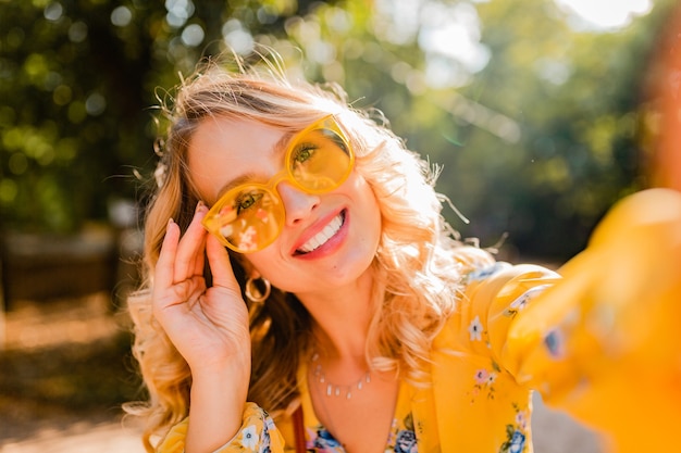 Ritratto di bella donna sorridente elegante bionda in camicetta gialla che indossa occhiali da sole facendo selfie foto