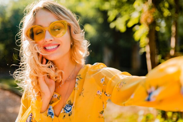 Ritratto di bella donna sorridente elegante bionda in camicetta gialla che indossa occhiali da sole facendo selfie foto