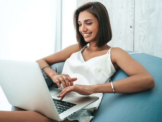 Ritratto di bella donna sorridente vestita di pigiama bianco. Modello spensierato seduto su una sedia morbida e usando il laptop.