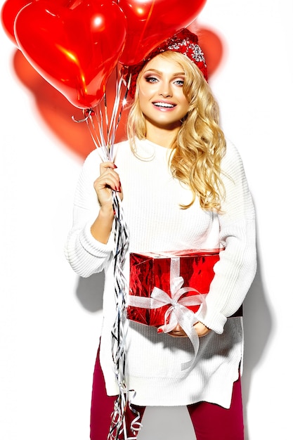 Ritratto di bella felice dolce donna bionda sorridente ragazza con in mano grande confezione regalo di Natale e palloncini a cuore in abiti invernali casual rosso hipster, in maglione bianco caldo
