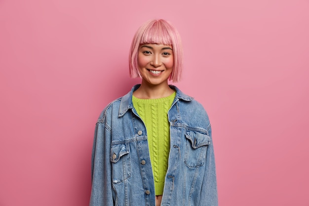 Ritratto di bella giovane donna con acconciatura rosa, sorride delicatamente, esprime emozioni positive, indossa un maglione corto verde, giacca di jeans, pose al coperto. Persone, giovani, concetto di sentimenti felici