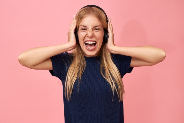 Ritratto di bella giovane donna con staffe di denti e capelli biondi in posa isolato in cuffie wireless, urlando, ascoltando musica tramite servizio di streaming online
