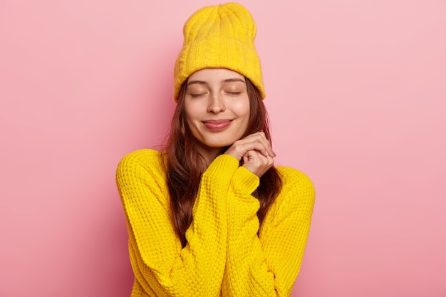 Ritratto di bella giovane donna europea tiene gli occhi chiusi, indossa un maglione lavorato a maglia giallo vivido e copricapo, isolato su sfondo rosa.