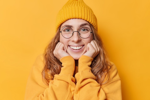 Ritratto di bella giovane donna europea tiene le mani sotto il mento sorride ampiamente mostra i denti bianchi focalizzati direttamente sulla fotocamera indossa una felpa casual e un cappello isolati su sfondo giallo studio