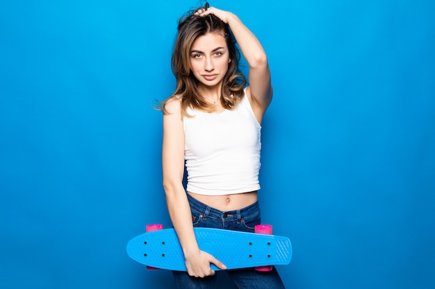 Ritratto di bella giovane donna in abiti casual in piedi, tenendo skateboard isolato sul muro blu. Concetto di lifestyle di persone.