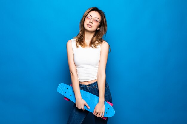 Ritratto di bella giovane donna in abiti casual in piedi, tenendo skateboard isolato sul muro blu. Concetto di lifestyle di persone.