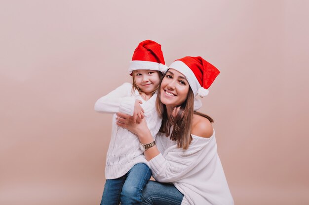 Ritratto di bella madre con piccola figlia carina che indossa pullover bianchi e cappelli di Babbo Natale