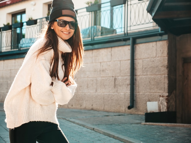 Ritratto di bella modella sorridente. Donna vestita di caldo maglione bianco hipster e berretto. Lei posa per strada