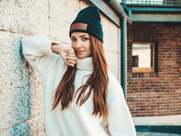 Ritratto di bella modella sorridente. Donna vestita di caldo maglione bianco hipster e berretto. Ragazza alla moda in posa vicino al muro in strada
