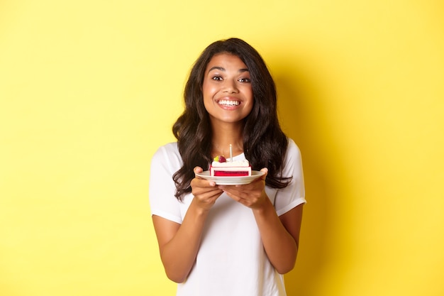 Ritratto di bella ragazza afro-americana che festeggia il compleanno, sorride e sembra felice e tiene in mano una torta di compleanno con una candela, in piedi su sfondo giallo