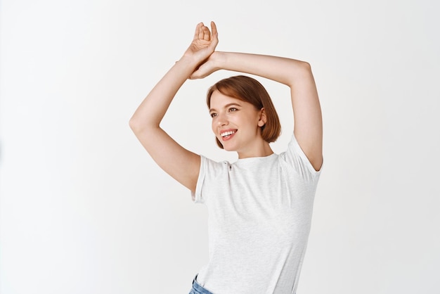 Ritratto di bella ragazza candida con trucco di luce naturale alzando le mani e guardando da parte con un sorriso spensierato in piedi in maglietta con jeans su sfondo bianco