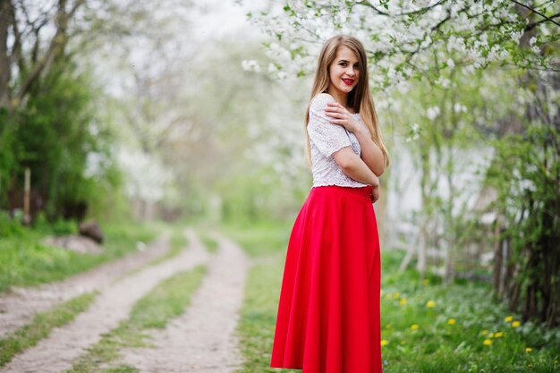 Ritratto di bella ragazza con le labbra rosse all'usura del giardino di fiori di primavera su abito rosso e camicetta bianca