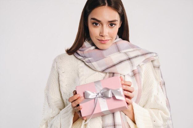 Ritratto di bella ragazza in maglione accogliente con sciarpa che tiene scatola regalo guardando attentamente nella fotocamera su sfondo bianco