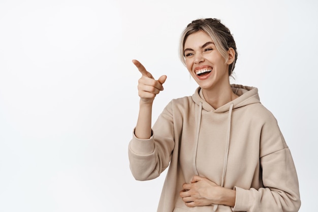 Ritratto di bella ragazza naturale che ride per qualcosa di divertente che punta il dito da parte in piedi in felpa con cappuccio beige su sfondo bianco