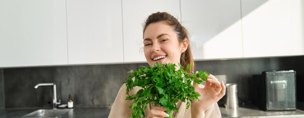 Ritratto di bella ragazza sorridente con il mazzo di prezzemolo in piedi in cucina e cucina