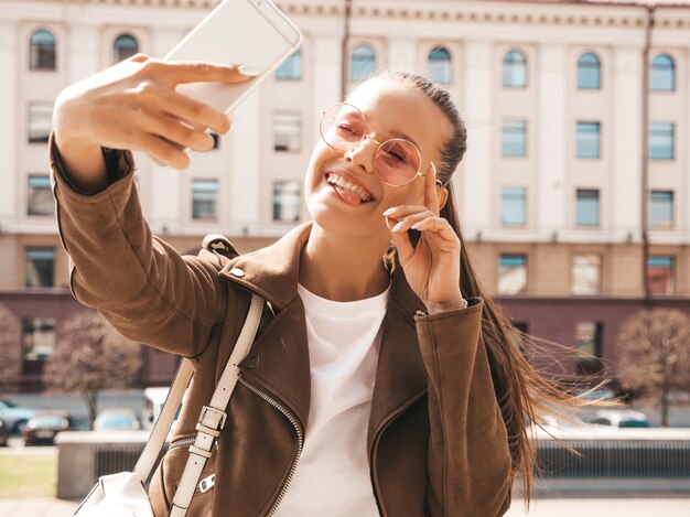 Ritratto di bella ragazza sorridente del brunette in rivestimento hipster estate. Selfie di presa di modello sullo smartphone. Mostrando la sua lingua